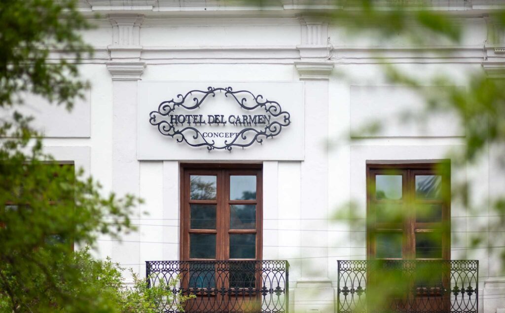 Fachada blanca con herrería y letrero de Hotel del Carmen entre árboles