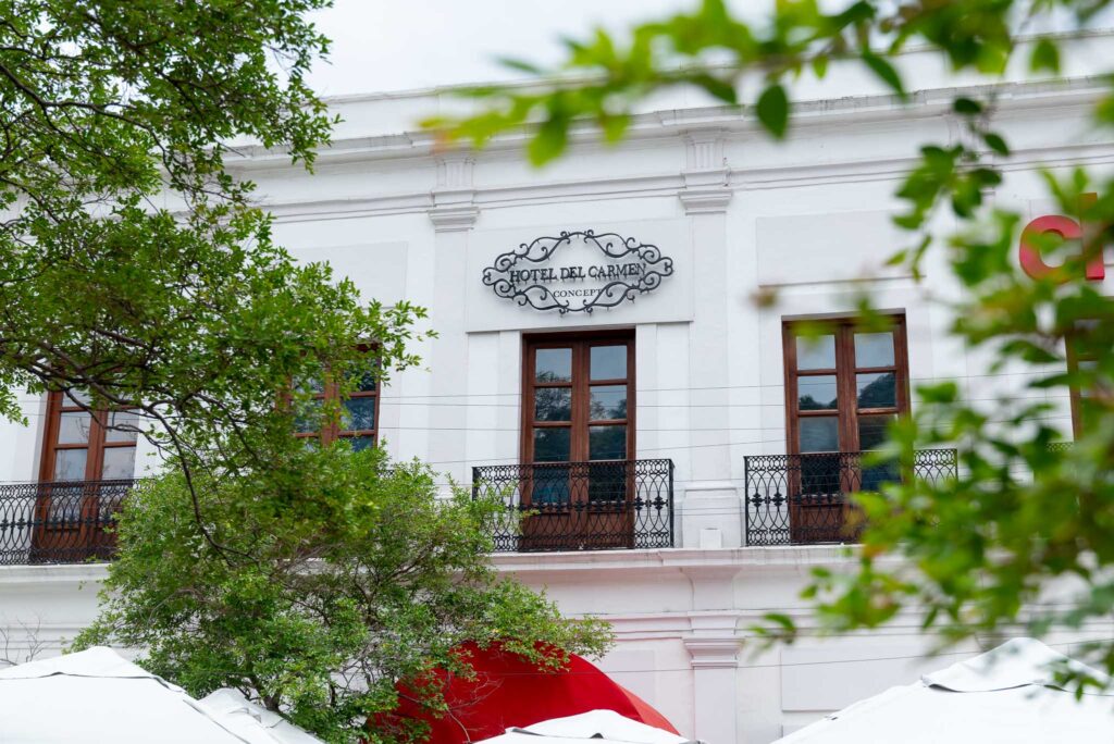 Fachada blanca con herrería y letrero de Hotel del Carmen entre árboles