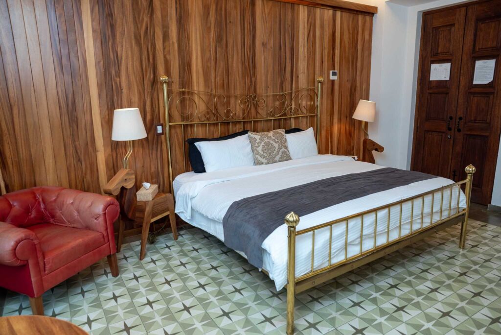Cama de latón con sillón de piel roja y muros de madera en la Suite Carrington